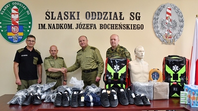 Funkcjonariusze Straży Granicznej przy przedmiotach przekazanych cudzoziemcom na tle napisu Śląski Oddział SG Funkcjonariusze Straży Granicznej przy przedmiotach przekazanych cudzoziemcom na tle napisu Śląski Oddział SG